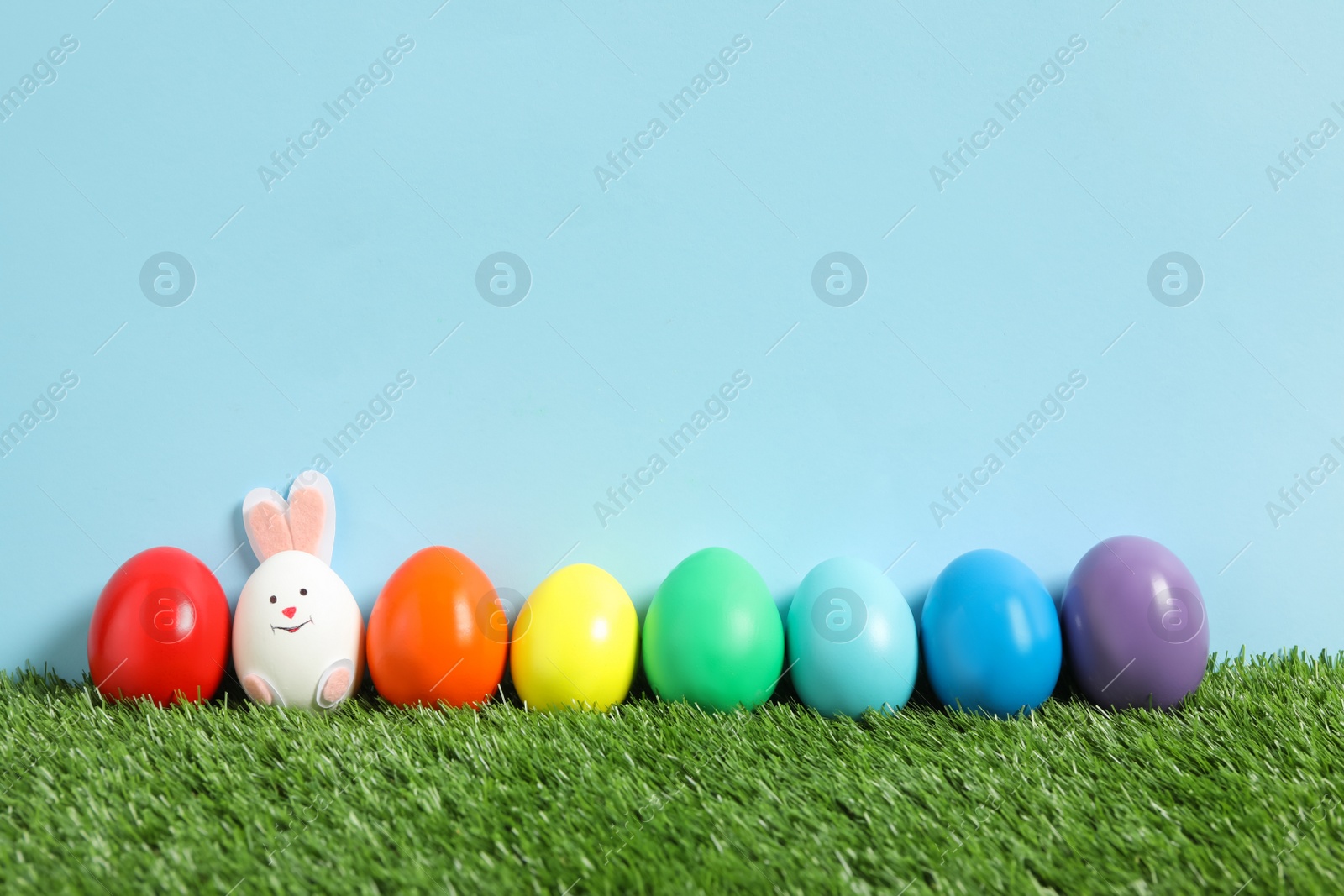 Photo of Bright Easter eggs with bunny ears on green grass against light blue background