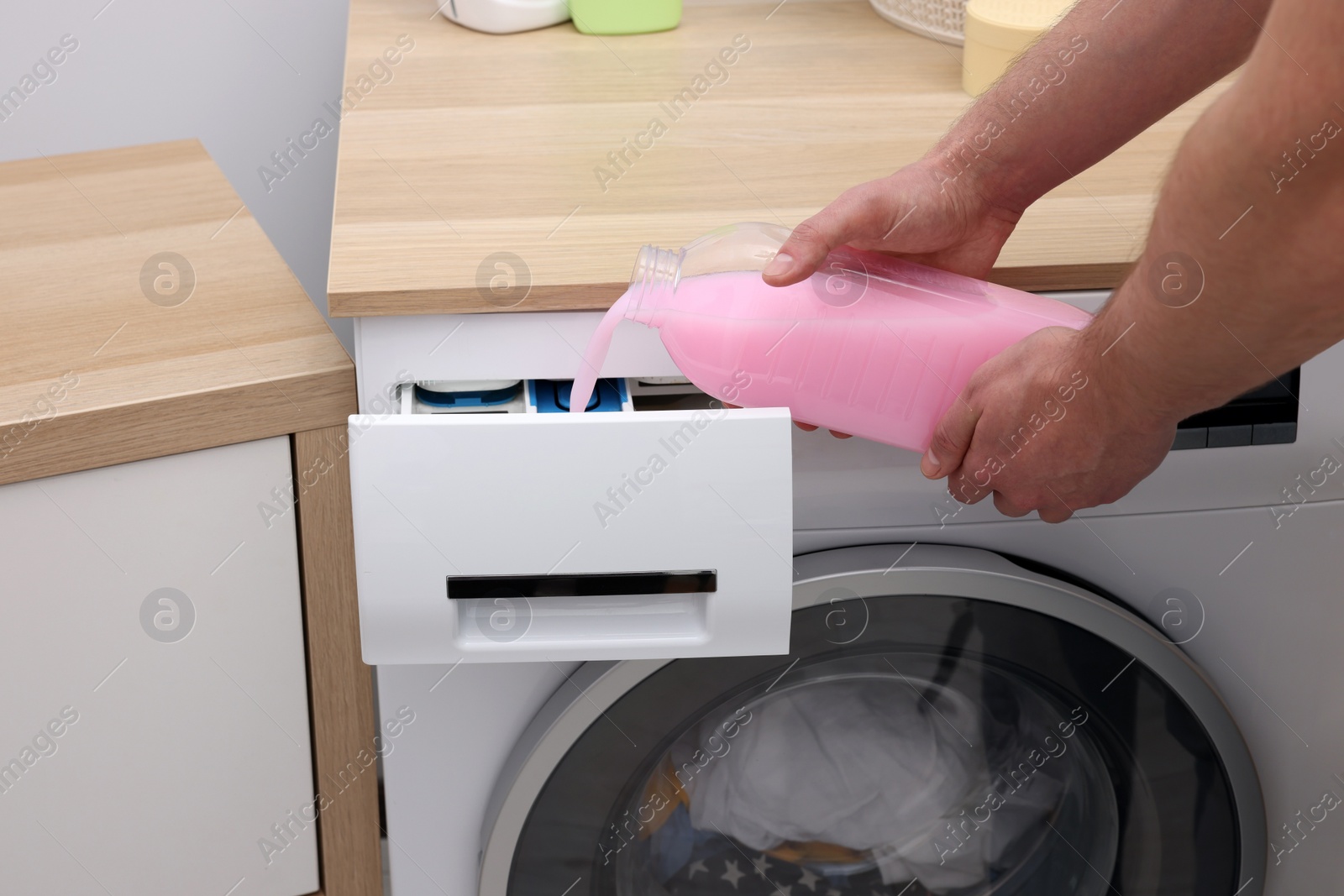 Photo of Man pouring fabric softener from bottle into washing machine indoors, closeup