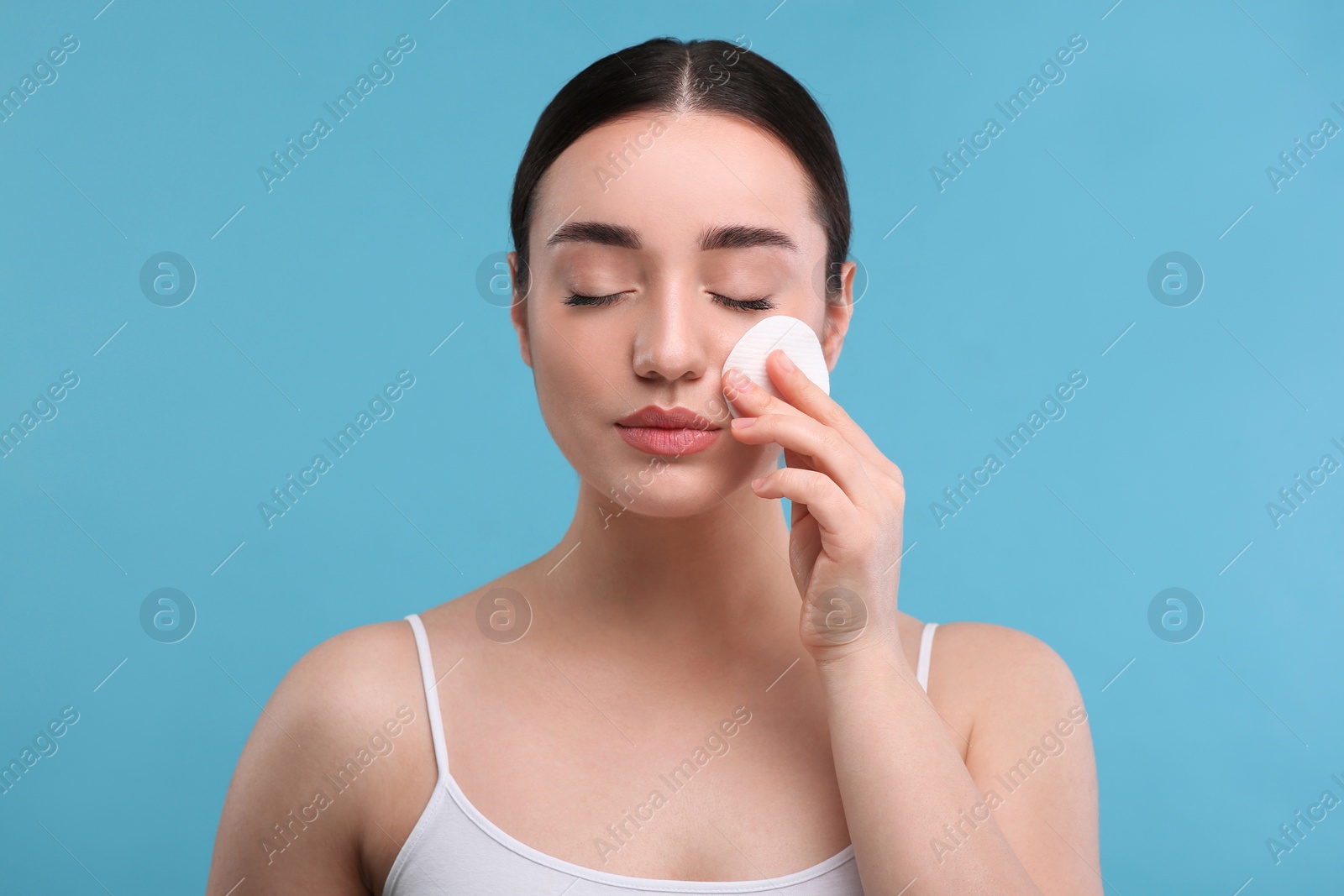 Photo of Beautiful woman removing makeup with cotton pad on light blue background