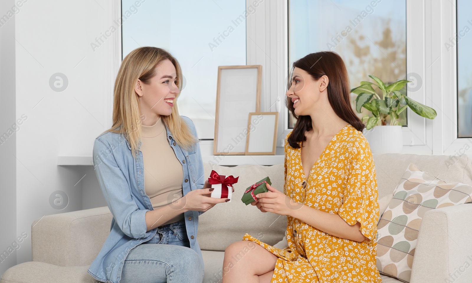 Photo of Smiling young women presenting gifts to each other on sofa at home