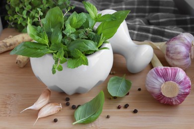 Mortar with different fresh herbs near garlic, horseradish roots and black peppercorns on wooden table, closeup