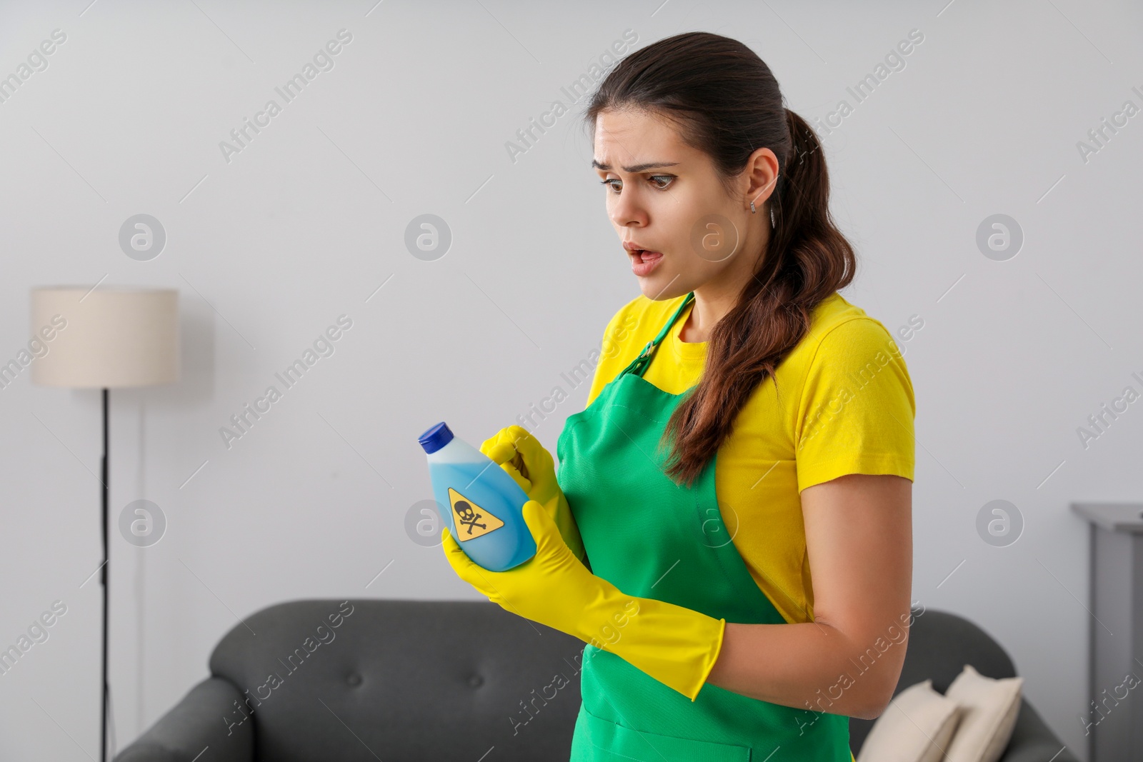 Photo of Woman looking at bottle of toxic household chemical with warning sign indoors