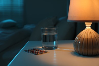 Photo of Insomnia treatment. Glass of water and pills on bedside table in bedroom at night