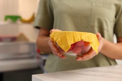 Woman holding bowl of fresh tomatoes covered with beeswax food wrap at table in kitchen, closeup. Space for text