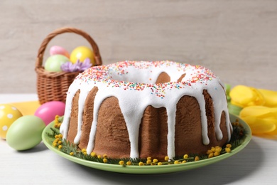 Photo of Glazed Easter cake with sprinkles, painted eggs and flowers on white wooden table