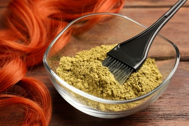 Photo of Bowl of henna powder, brush and red strand on wooden table, closeup. Natural hair coloring