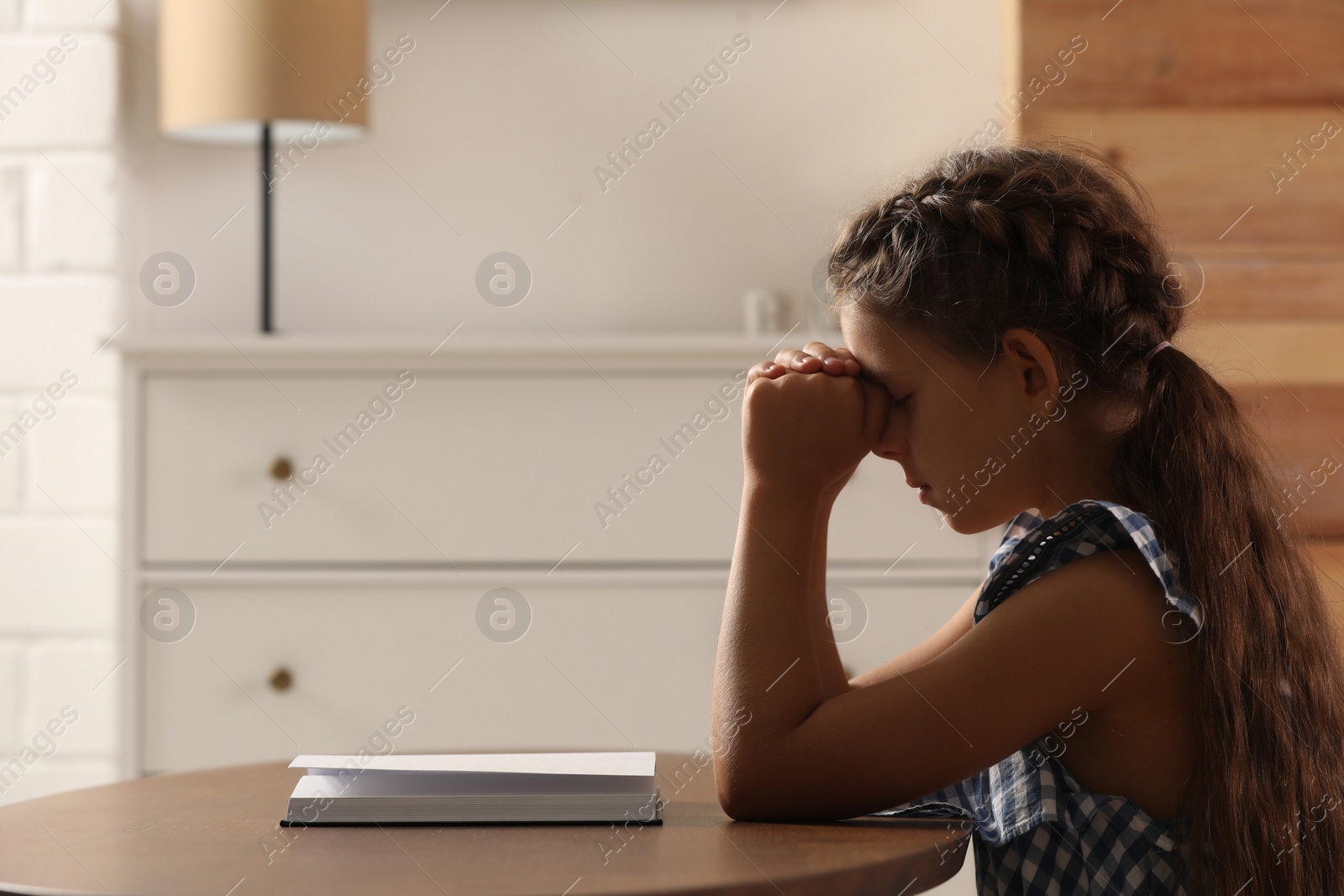 Photo of Cute little girl praying over Bible at table in room. Space for text