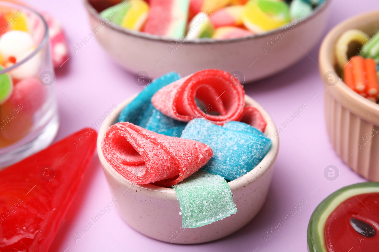 Photo of Bowl with different jelly candies on lilac background, closeup