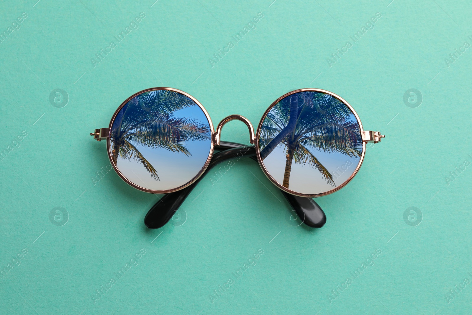 Image of Elegant sunglasses on turquoise background, top view. Sky and palm trees reflecting in lenses