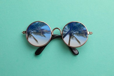 Image of Elegant sunglasses on turquoise background, top view. Sky and palm trees reflecting in lenses