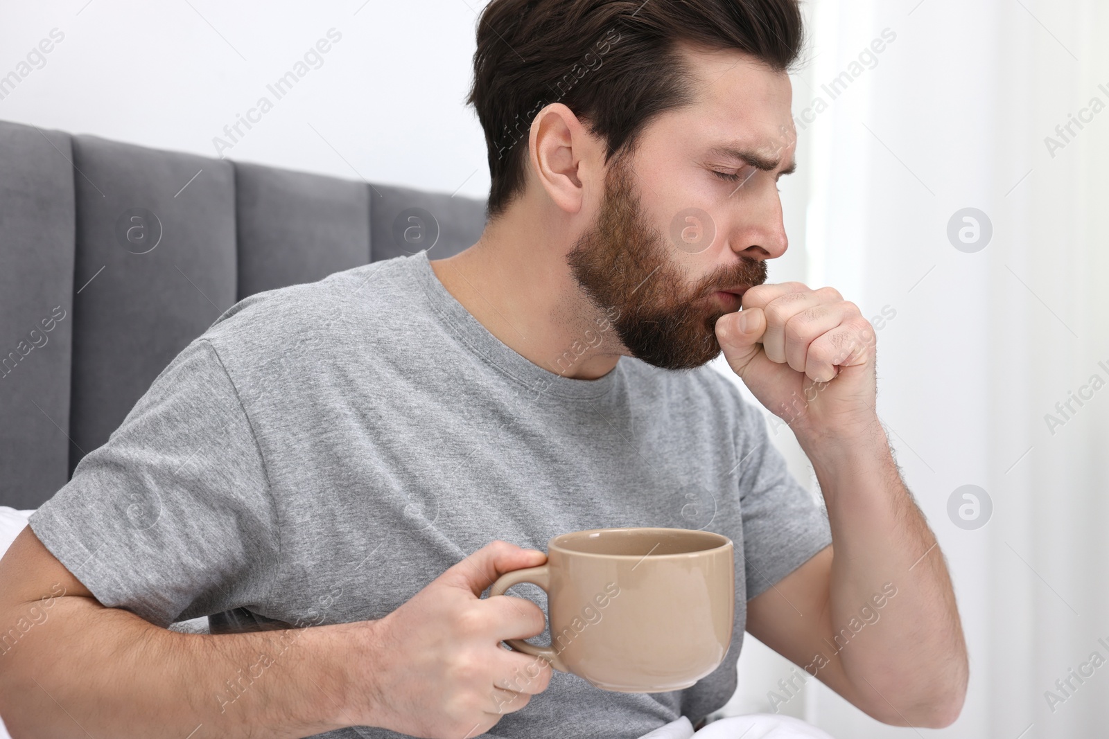 Photo of Sick man with cup of hot drink coughing on bed at home. Cold symptoms