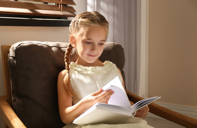 Photo of Little girl reading book in armchair at home