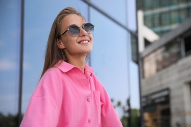 Portrait of beautiful young woman in stylish sunglasses near building outdoors, space for text