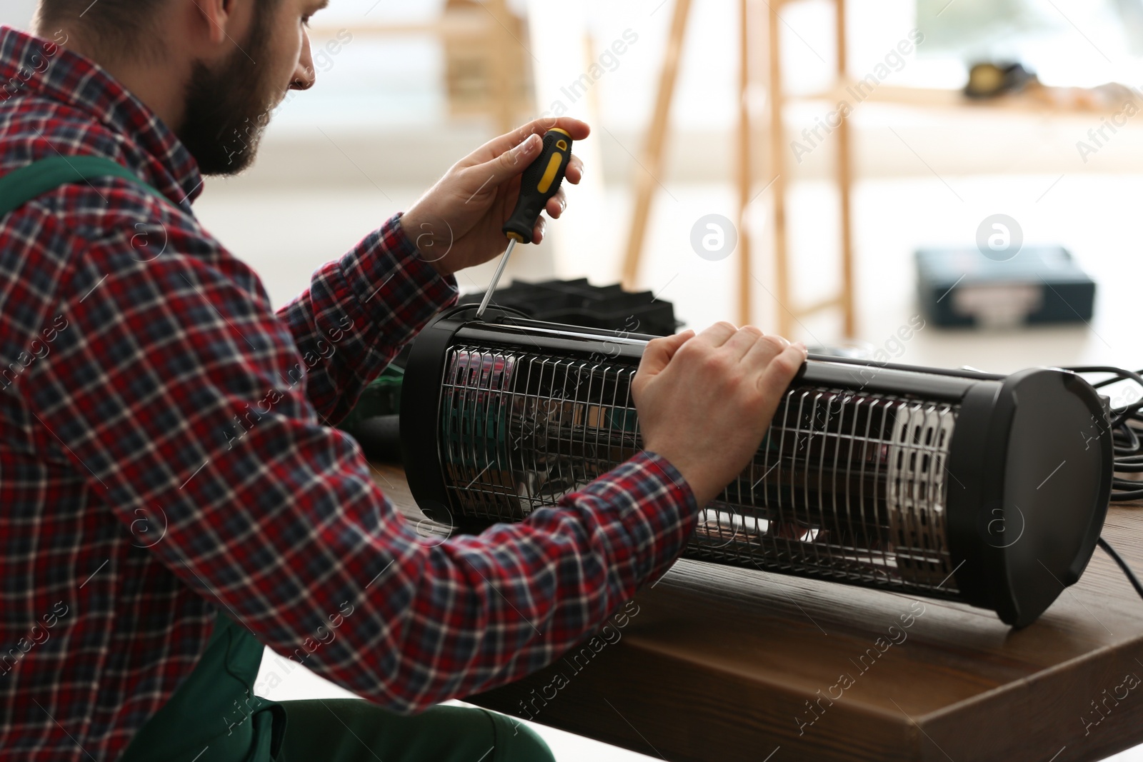 Photo of Professional technician repairing electric halogen heater with screwdriver at table indoors, closeup