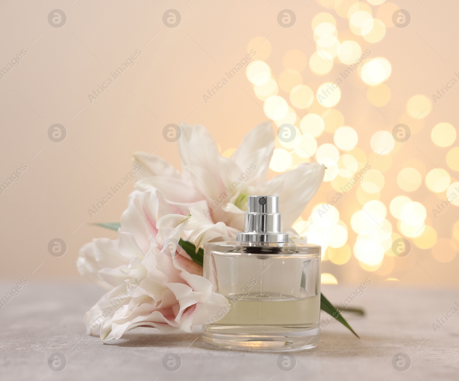 Photo of Bottle of perfume and beautiful lily flowers on table against beige background with blurred lights