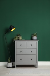 Modern chest of drawers with houseplants and lamp near green wall indoors