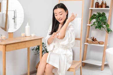 Photo of Beautiful young Asian woman applying body cream on elbow in bathroom