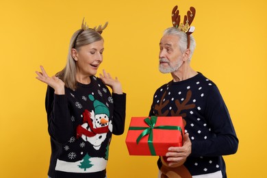 Photo of Couple in Christmas sweaters. Senior man presenting gift to his woman on orange background