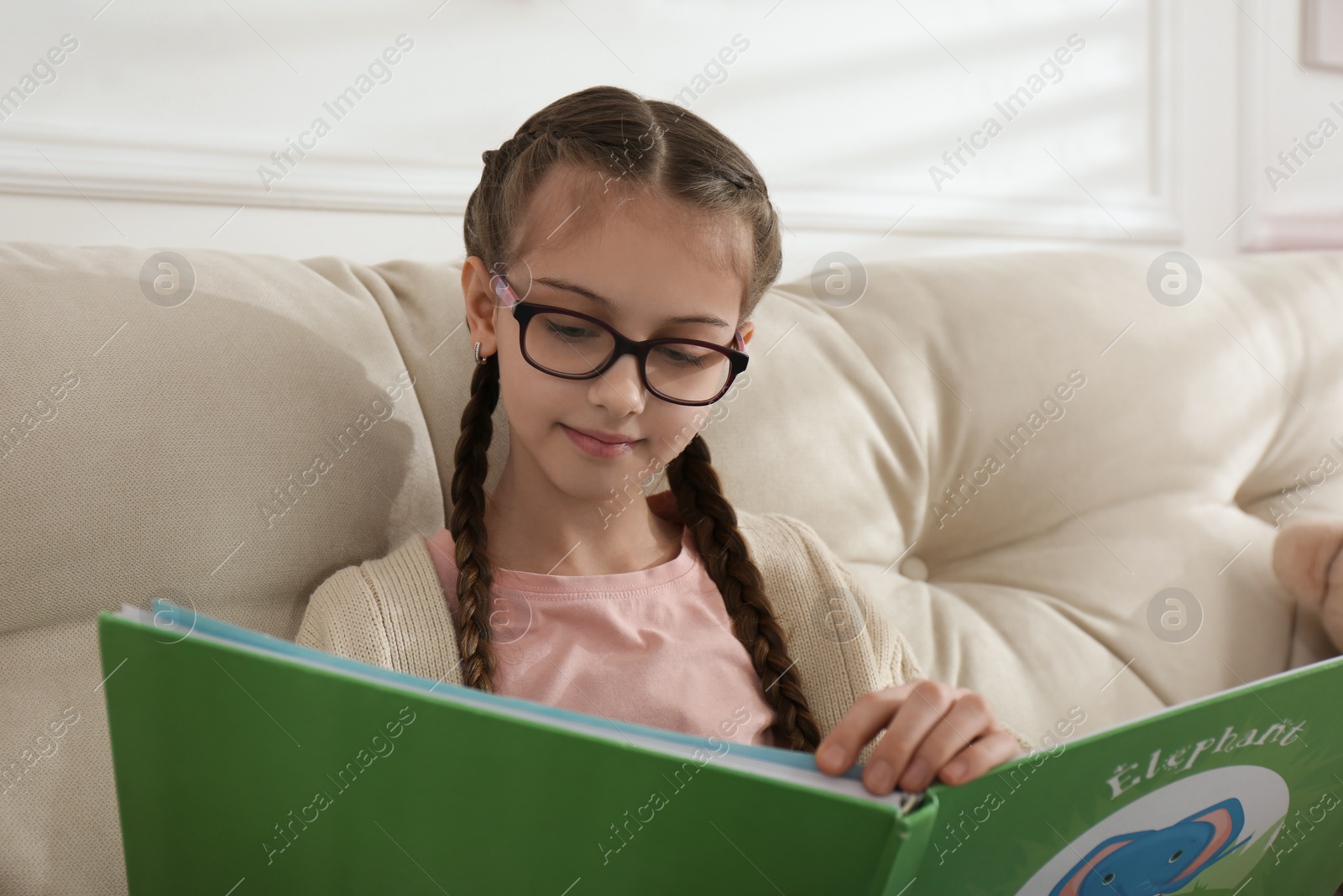 Photo of Cute little girl reading book on sofa at home