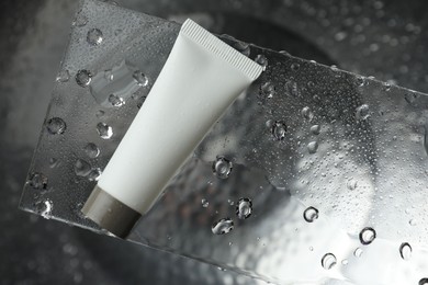Photo of Moisturizing cream in tube on glass with water drops against metal background, top view