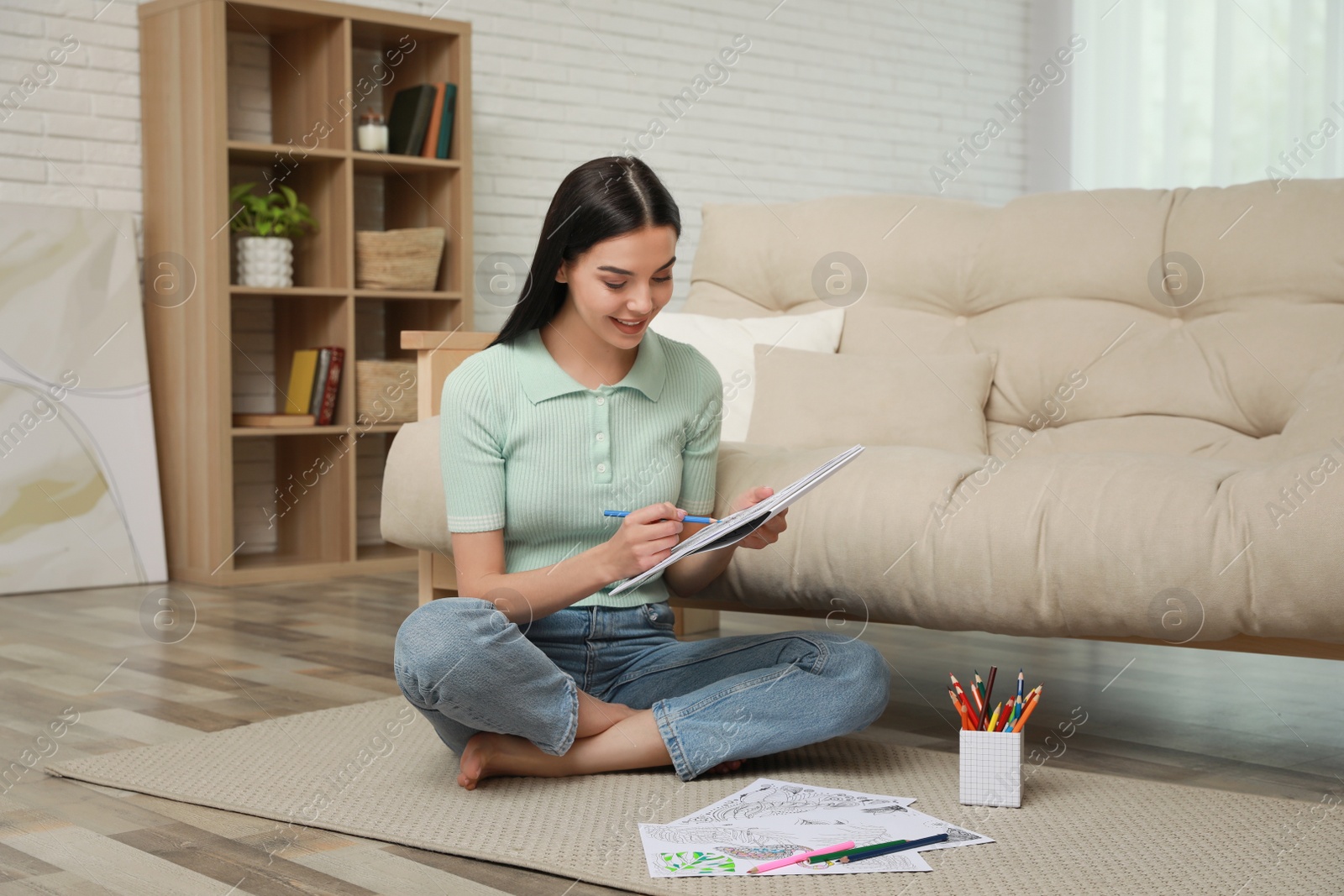 Photo of Young woman coloring antistress page near sofa in living room