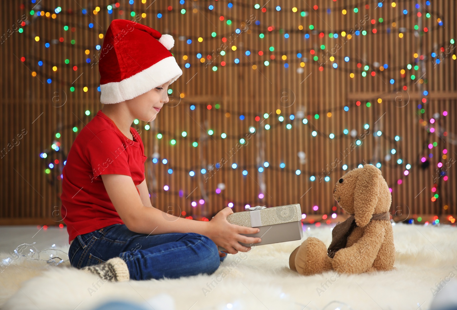 Photo of Cute little child in Santa hat giving Christmas gift box to toy rabbit against blurred lights