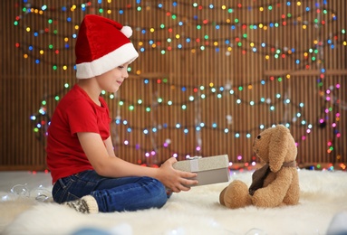 Cute little child in Santa hat giving Christmas gift box to toy rabbit against blurred lights