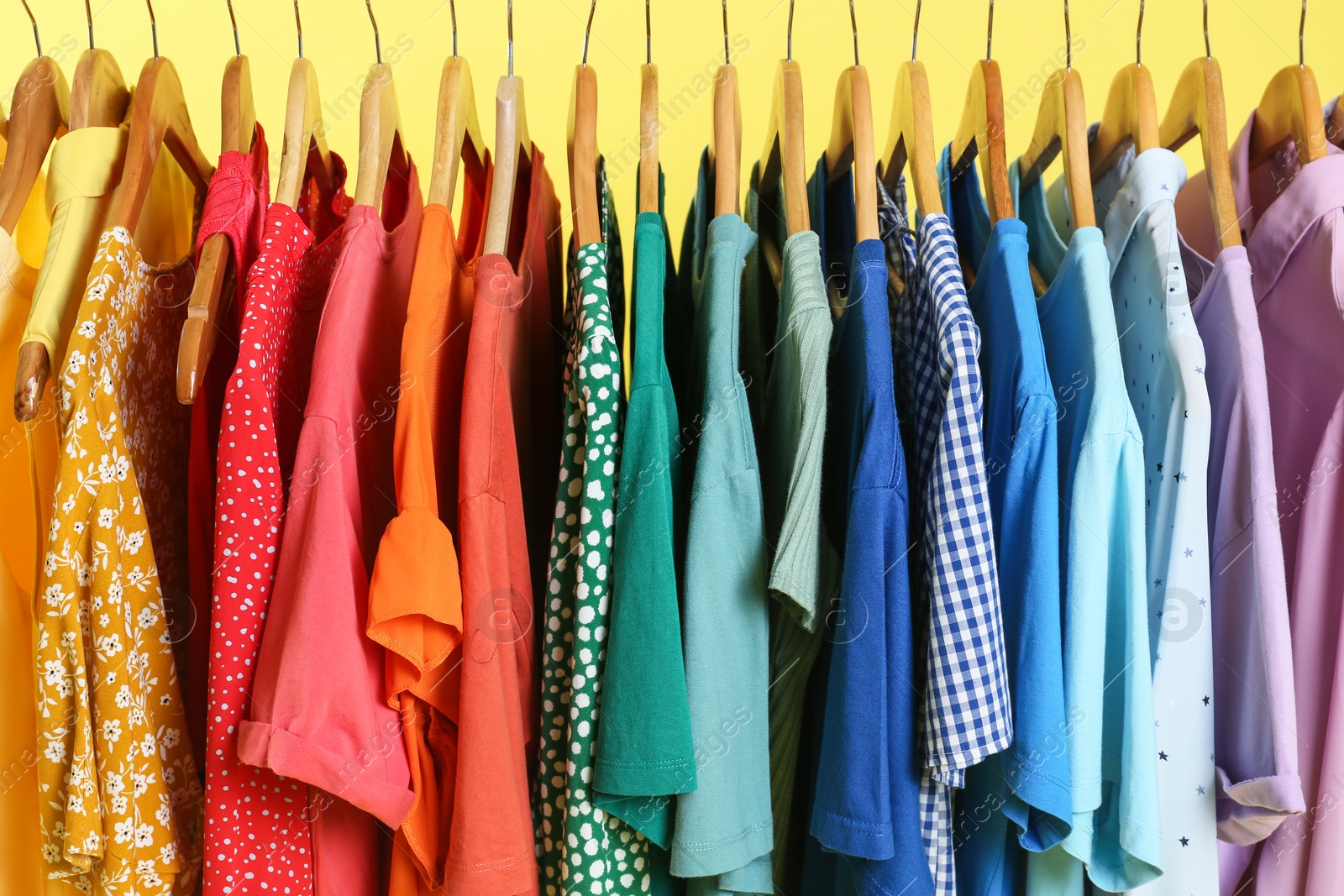 Photo of Bright clothes on hangers against yellow background, closeup. Rainbow colors