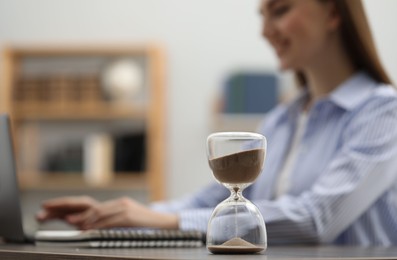 Photo of Hourglass with flowing sand on desk. Woman using laptop indoors, selective focus