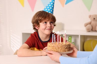 Birthday celebration. Mother holding tasty cake with burning candles near her son indoors