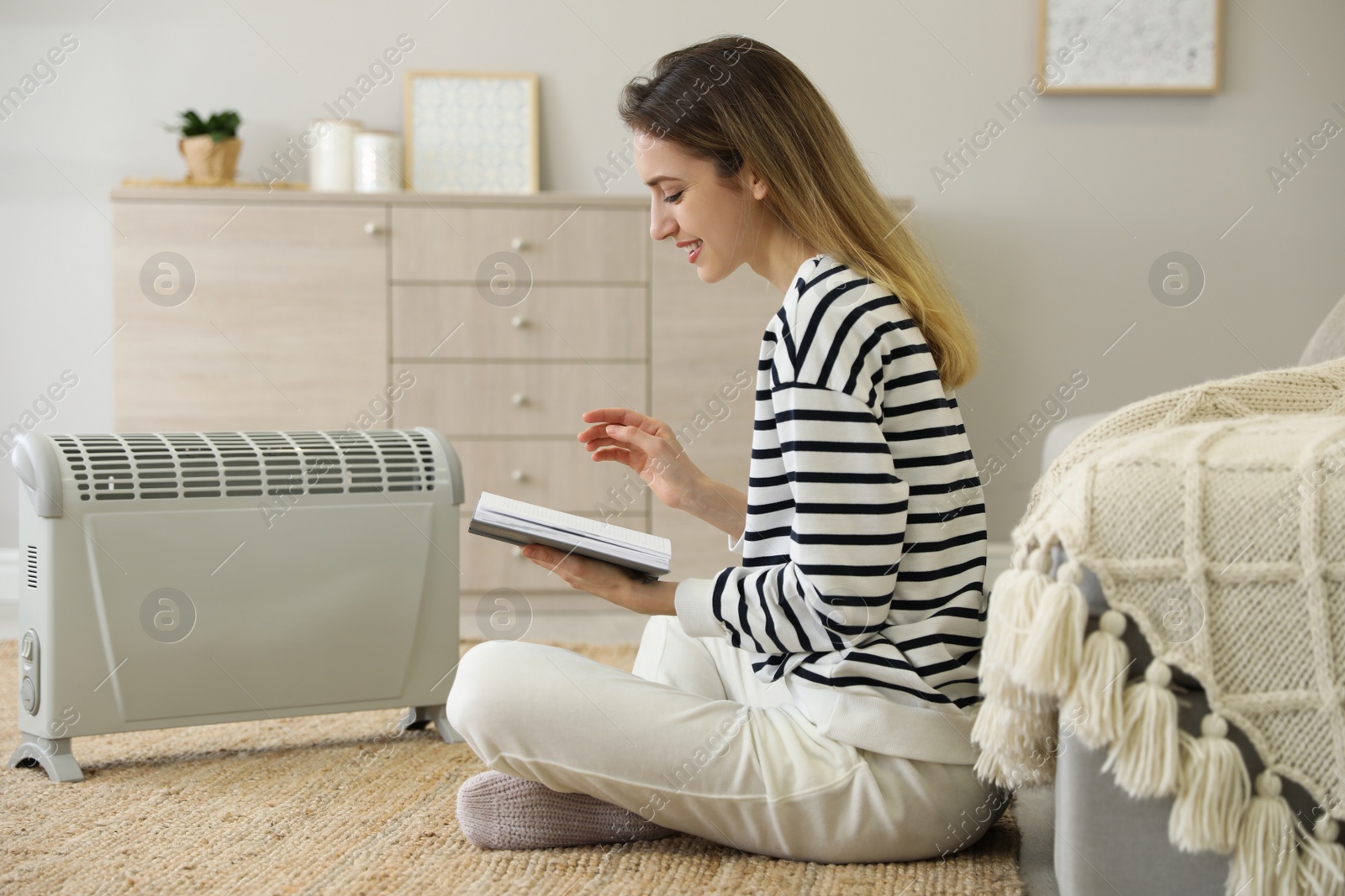 Photo of Young woman with book sitting on floor near electric heater at home