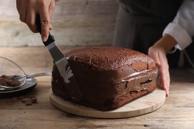 Photo of Woman smearing sides of sponge cake with chocolate cream at wooden table, closeup