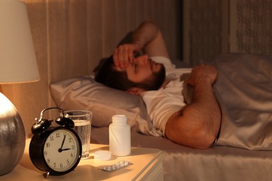 Photo of Man suffering from insomnia in bed at night, focus on nightstand with pills and alarm clock