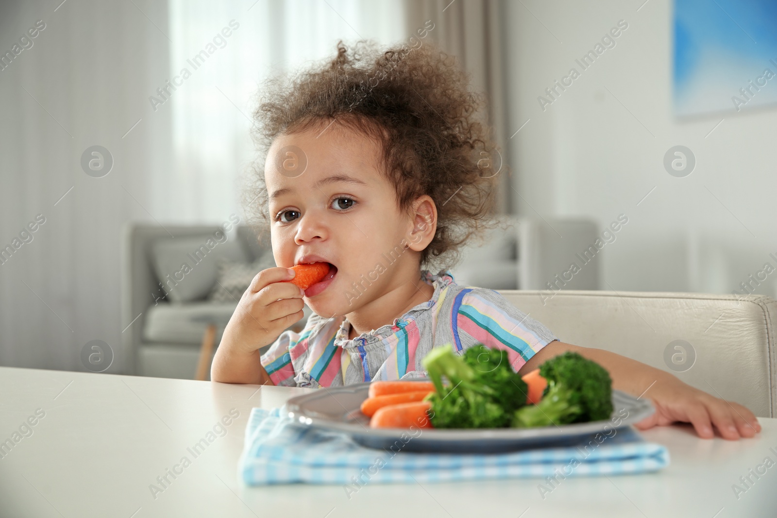 Photo of Cute African-American girl eating vegetables at table in living room