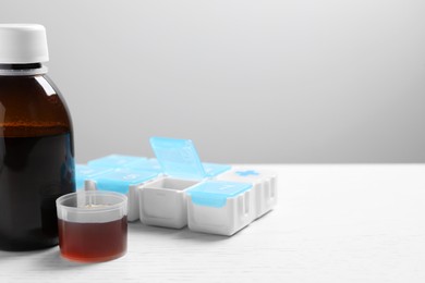 Photo of Bottle of syrup, measuring cup and weekly pill organizer on white table against light grey background, space for text. Cold medicine