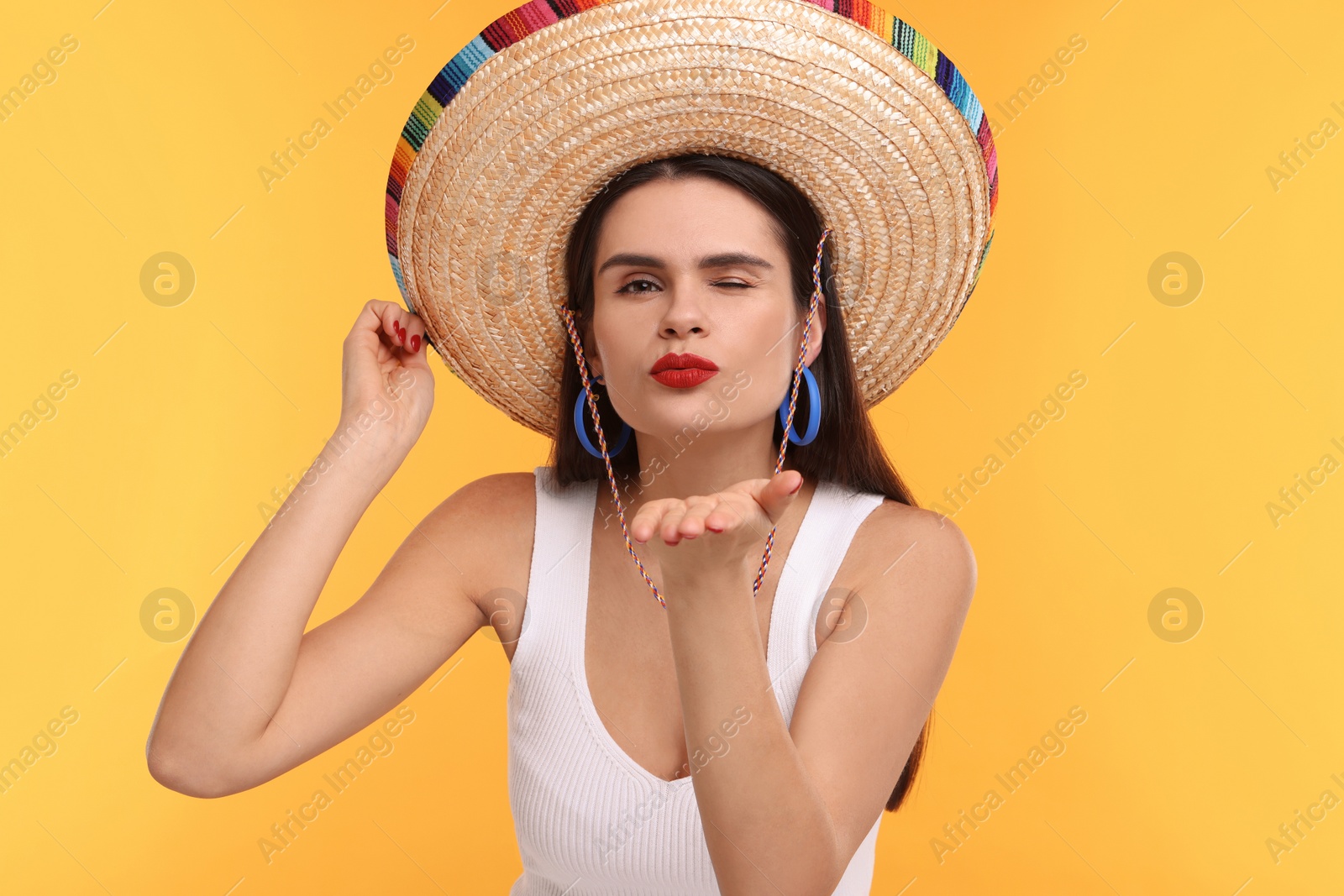 Photo of Young woman in Mexican sombrero hat blowing kiss on yellow background