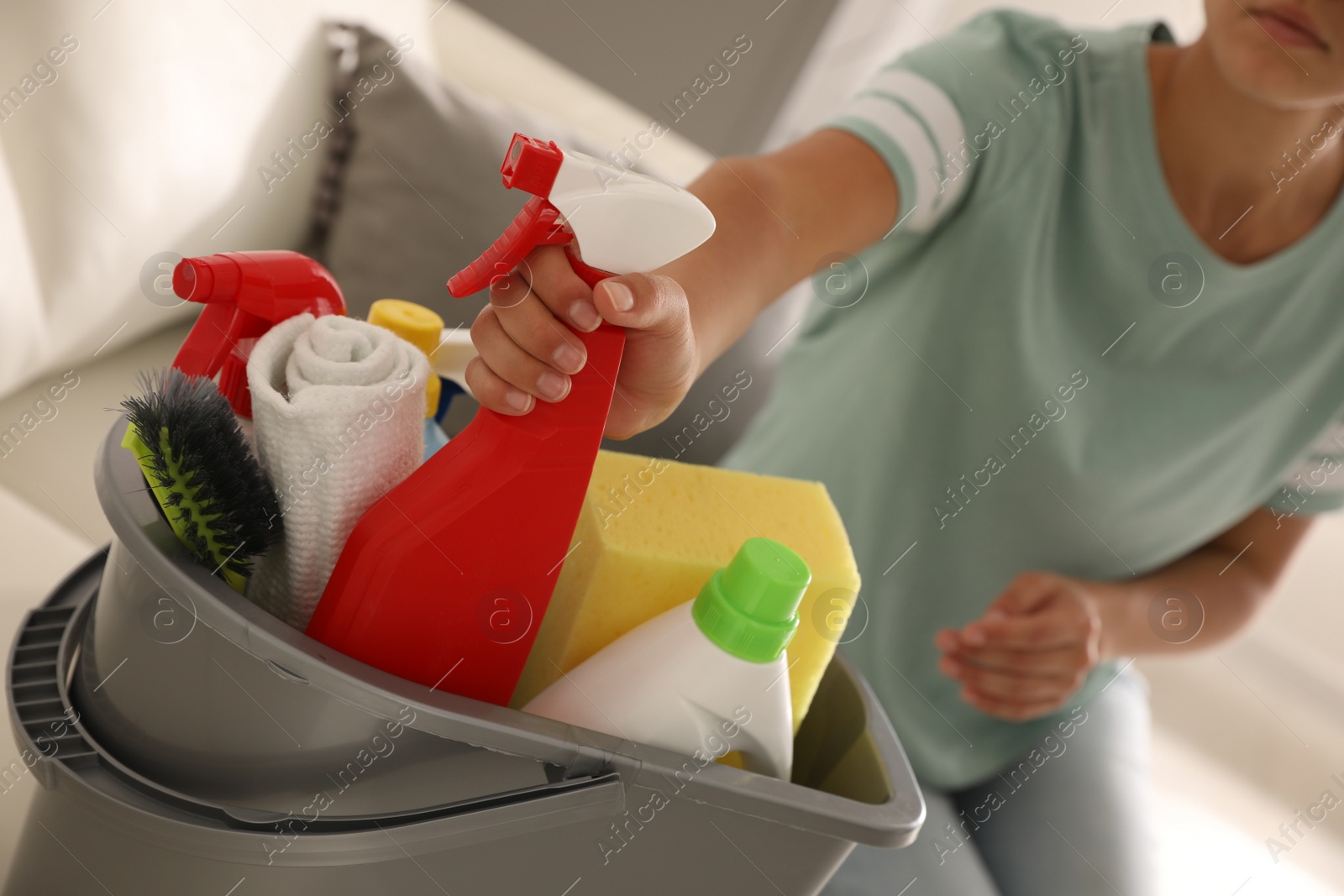 Photo of Woman taking bottle of detergent from bucket with cleaning supplies indoors, closeup