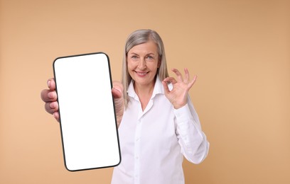 Happy mature woman showing mobile phone with blank screen on beige background. Mockup for design