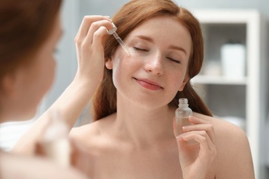 Beautiful woman with freckles applying cosmetic serum onto her face near mirror in bathroom