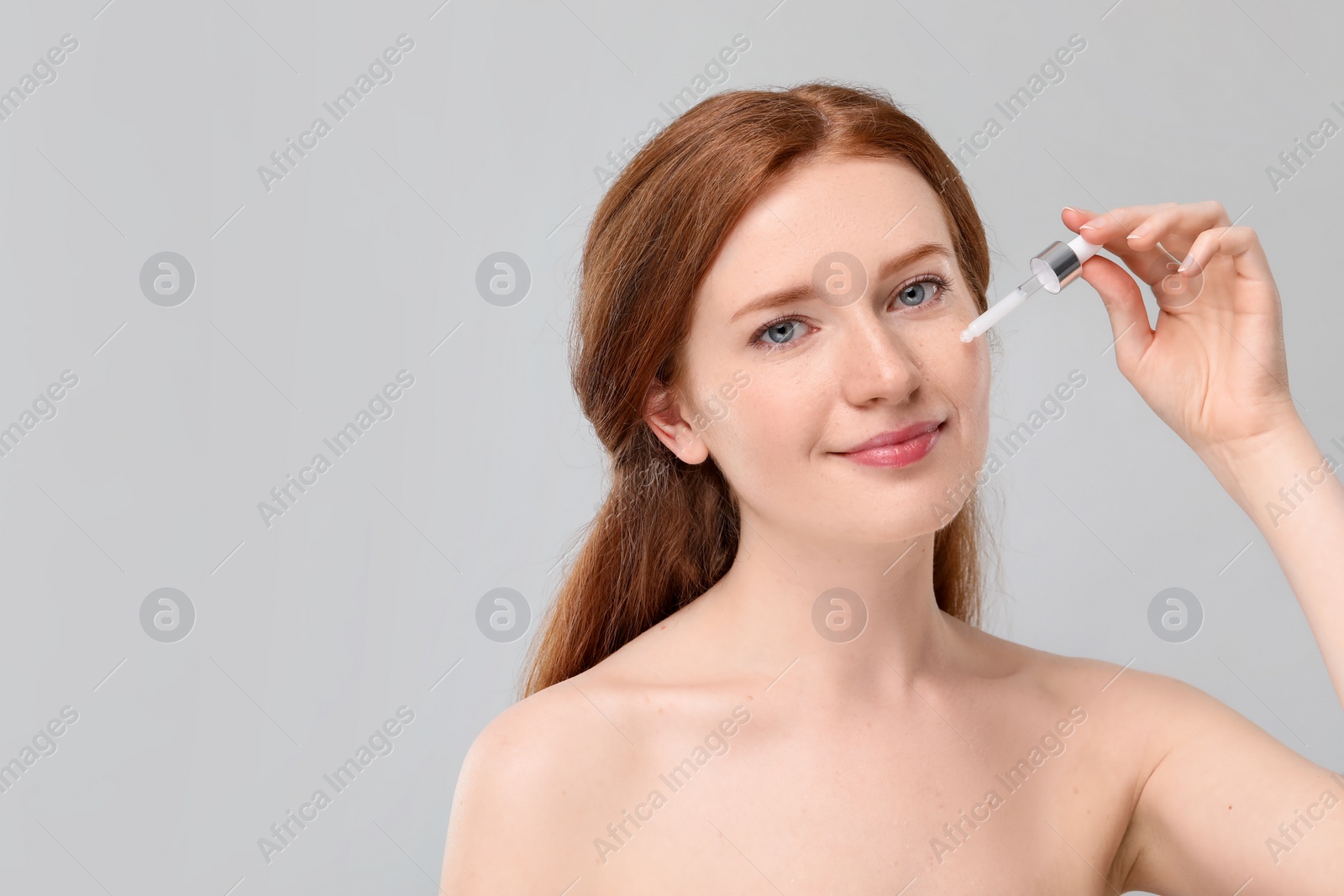 Photo of Beautiful woman with freckles applying cosmetic serum onto her face against grey background. Space for text