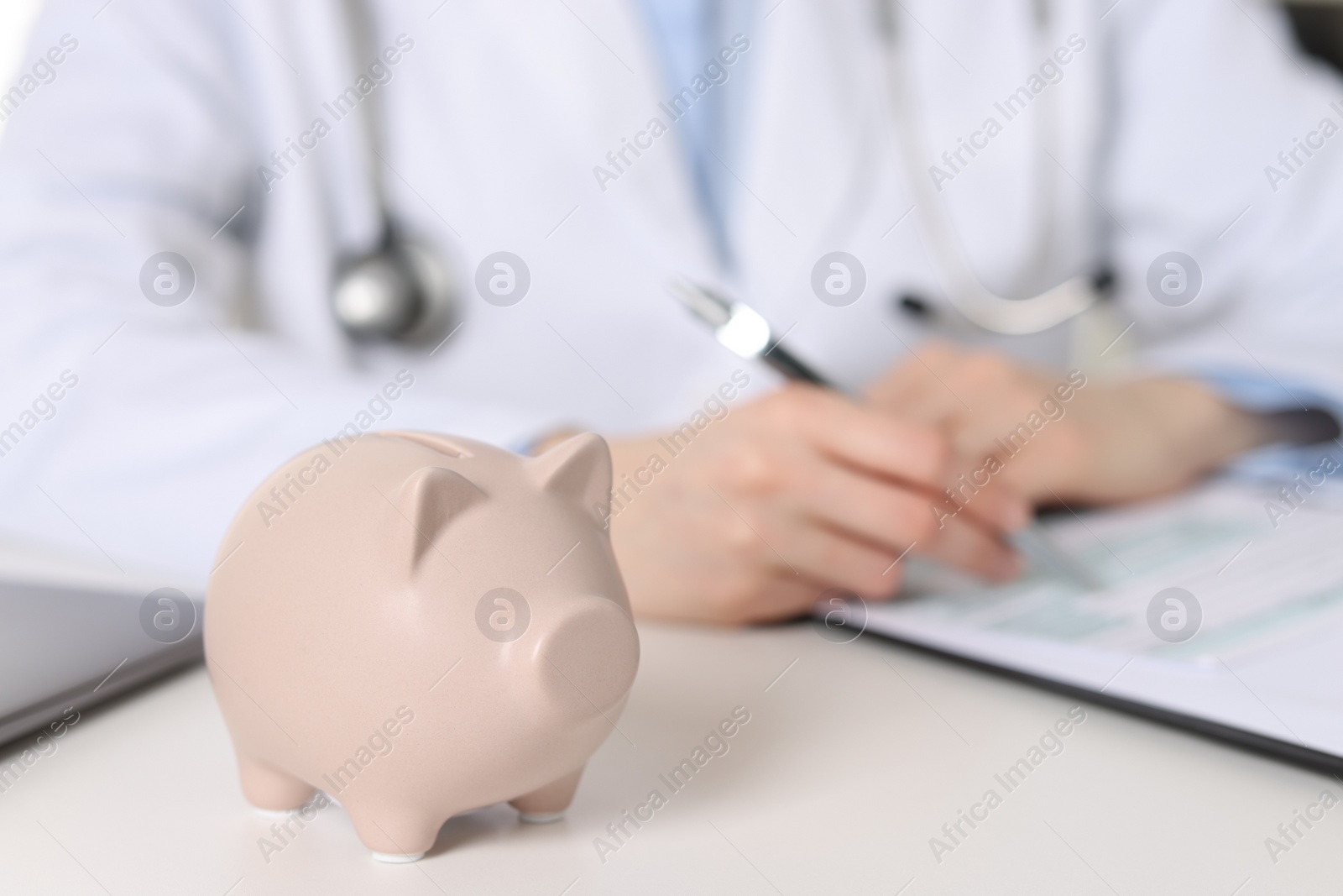 Photo of Doctor making notes at white table, focus on piggy bank