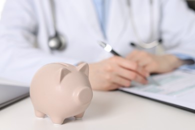 Photo of Doctor making notes at white table, focus on piggy bank