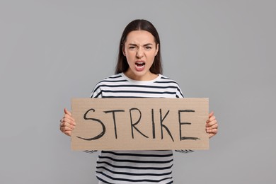 Angry young woman holding cardboard banner with word Strike on light grey background