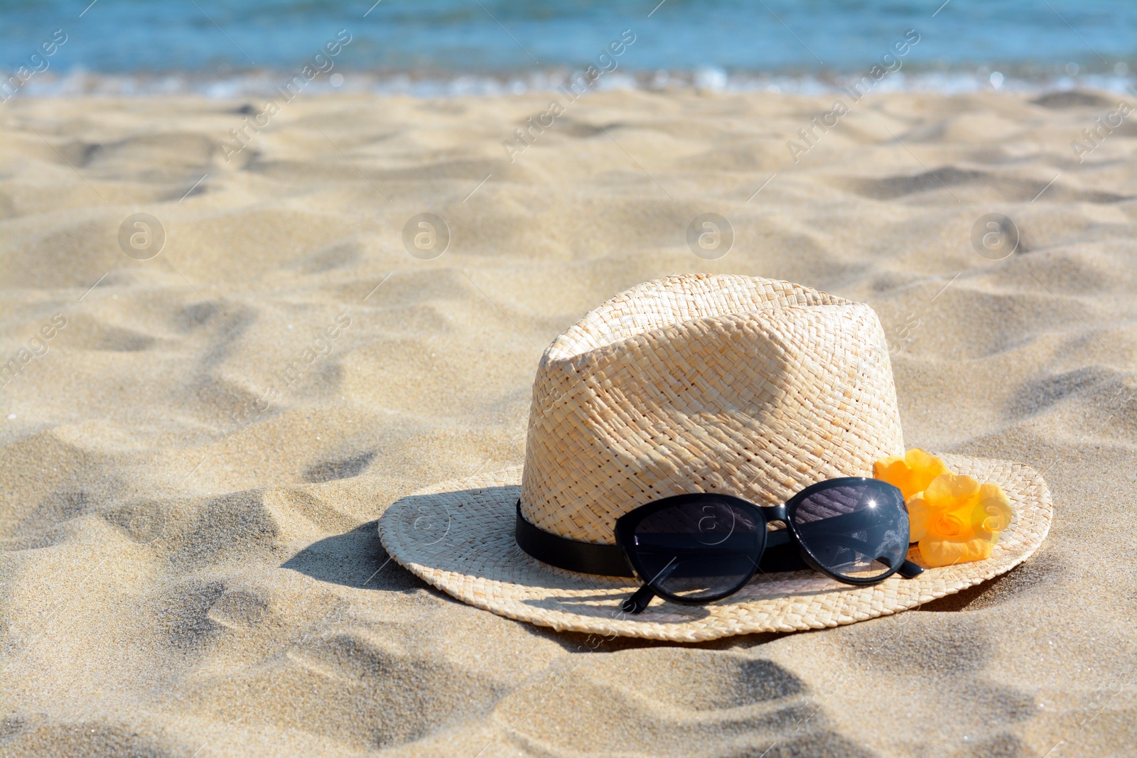 Photo of Hat with beautiful sunglasses and flowers on sand near sea. Space for text