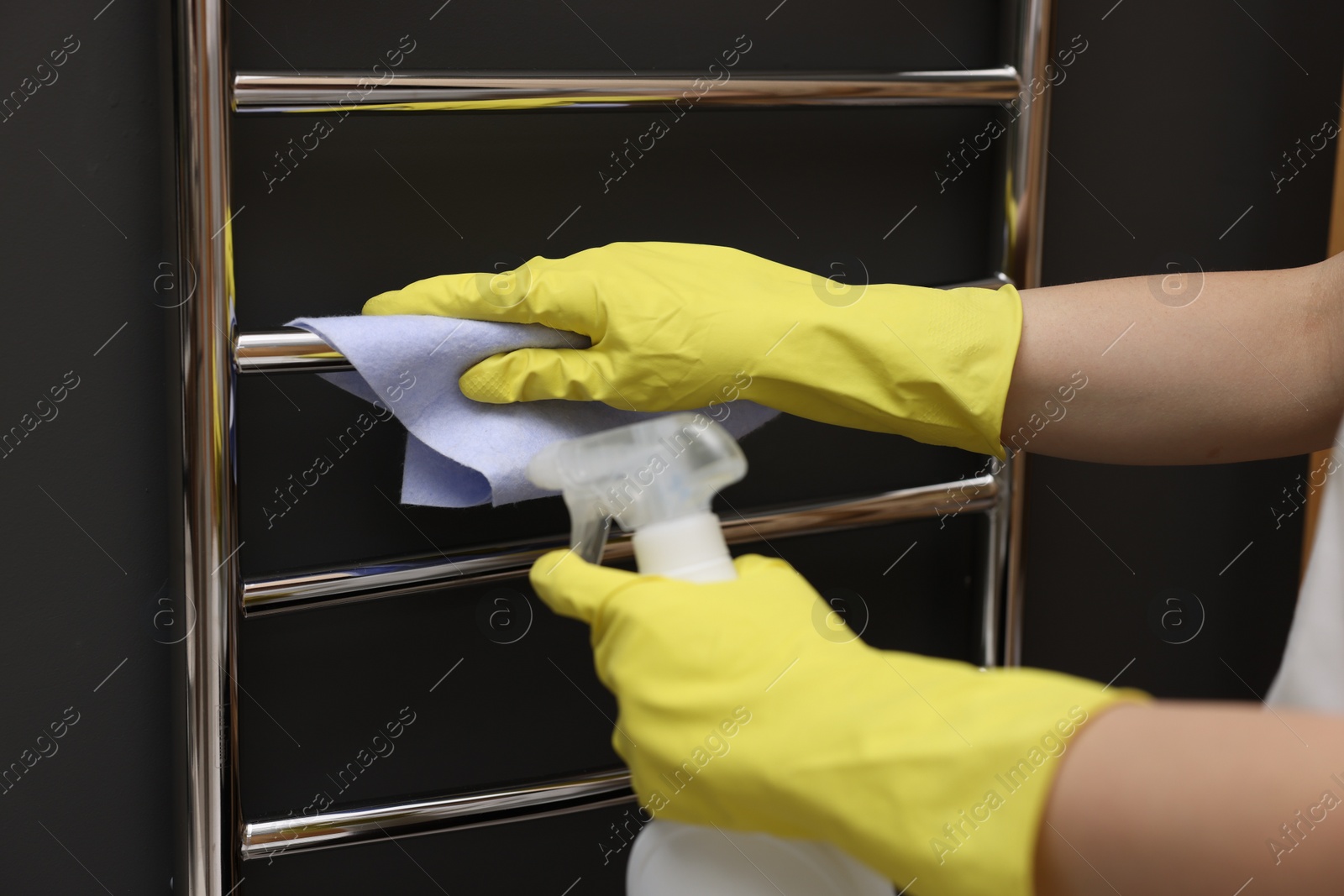 Photo of Woman cleaning heated towel rail with sprayer and rag, closeup