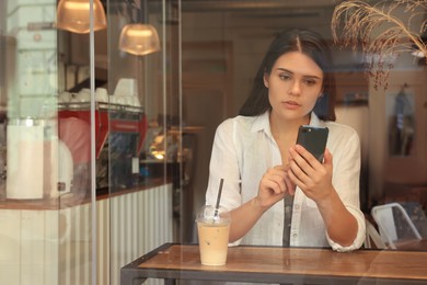 Photo of Young beautiful woman with smartphone in cafe, view through window glass