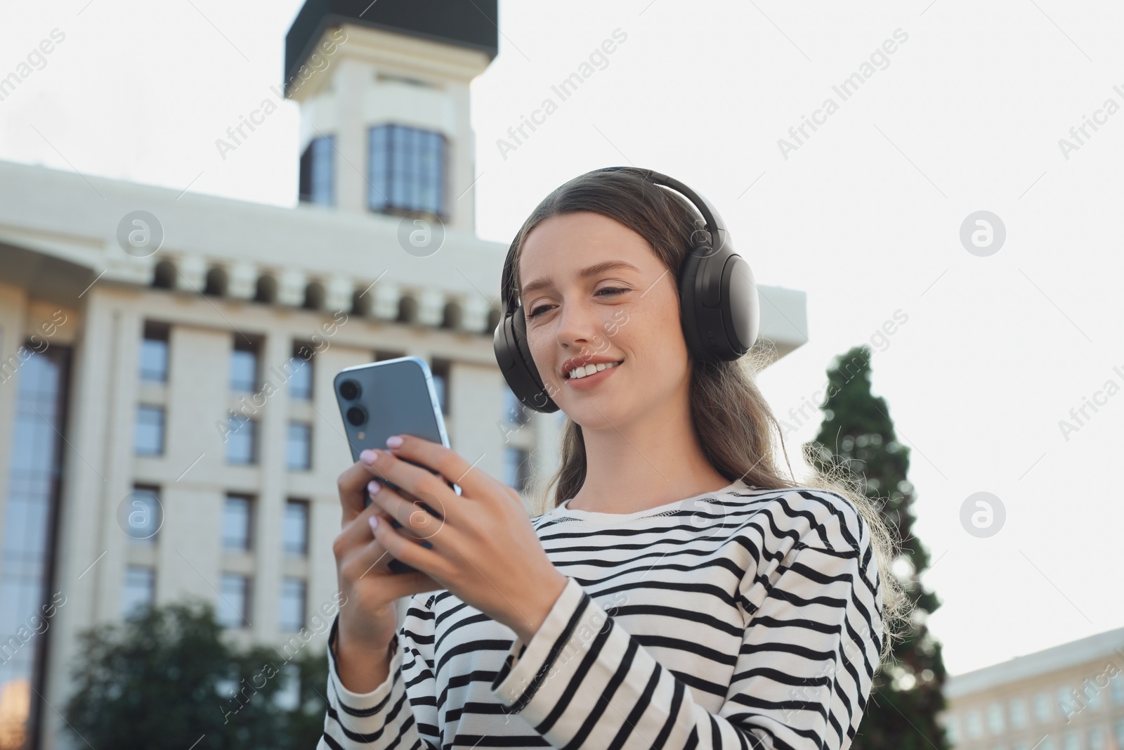 Photo of Smiling woman in headphones using smartphone on city street