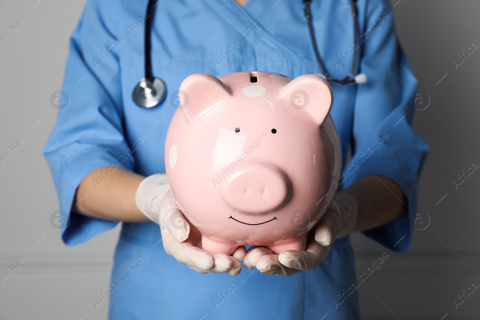Photo of Doctor holding pale pink ceramic piggy bank against white wall, closeup. Medical insurance