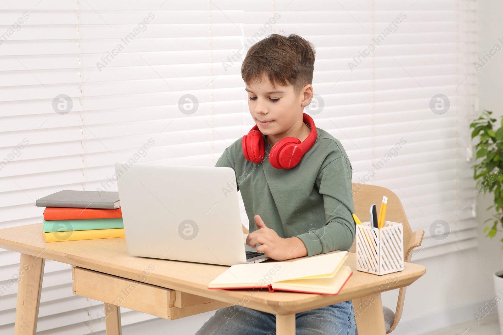 Photo of Boy with red headphones using laptop at desk in room. Home workplace