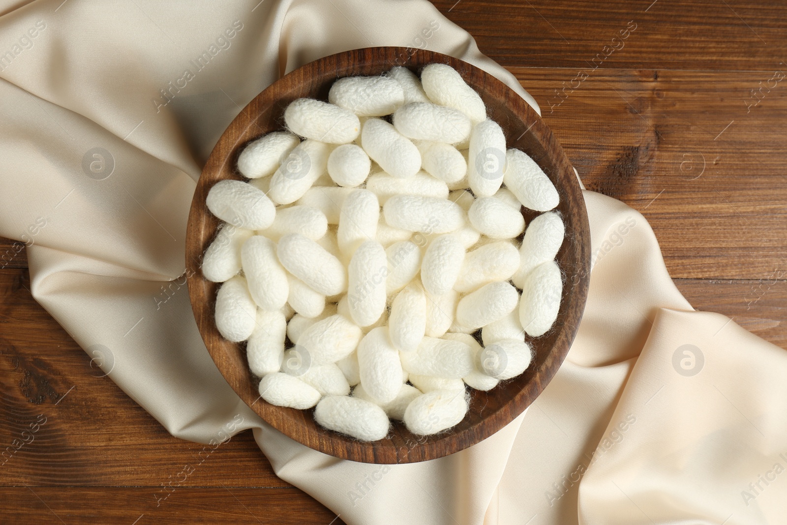 Photo of White cocoons with bowl and silk fabric on wooden table, flat lay